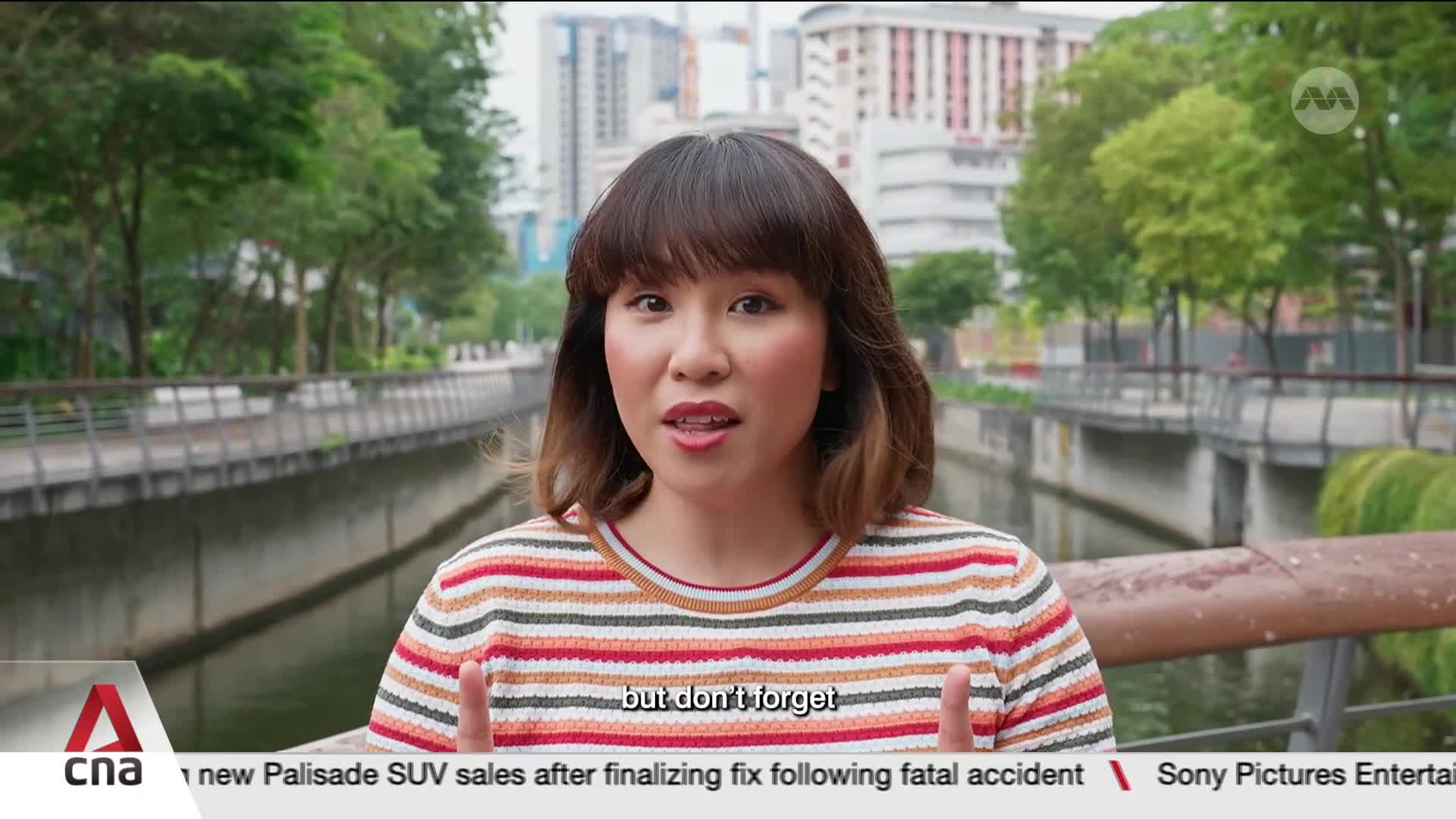 A woman in a striped shirt speaks directly to me, her hands gesturing slightly. Behind her, the Singapore River flows calmly, bordered by trees and modern buildings.