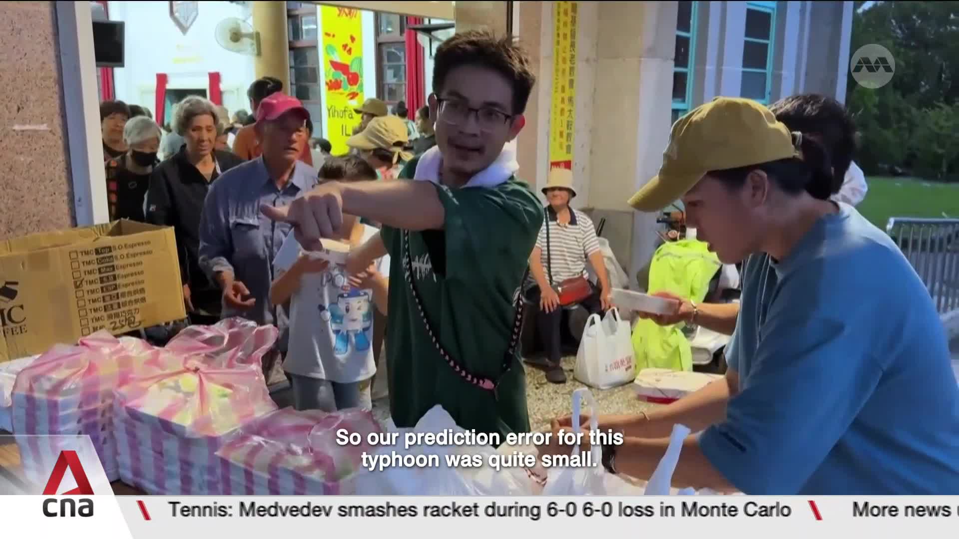 Volunteers hand out food packages to a line of people. A man in a green shirt gestures while speaking, his words about a typhoon prediction appearing on screen.