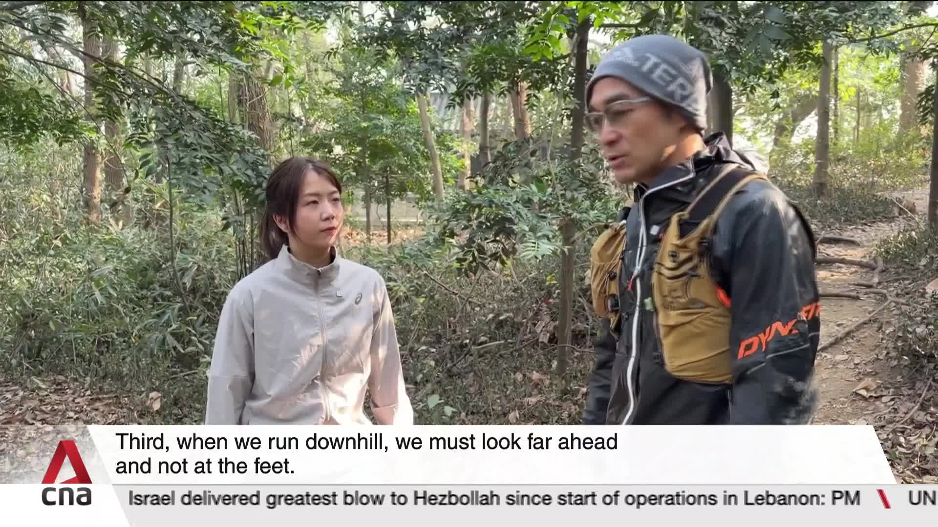 A man in a running vest and beanie talks to a woman in a light jacket on a wooded trail. The path ahead dips downhill, surrounded by lush greenery.