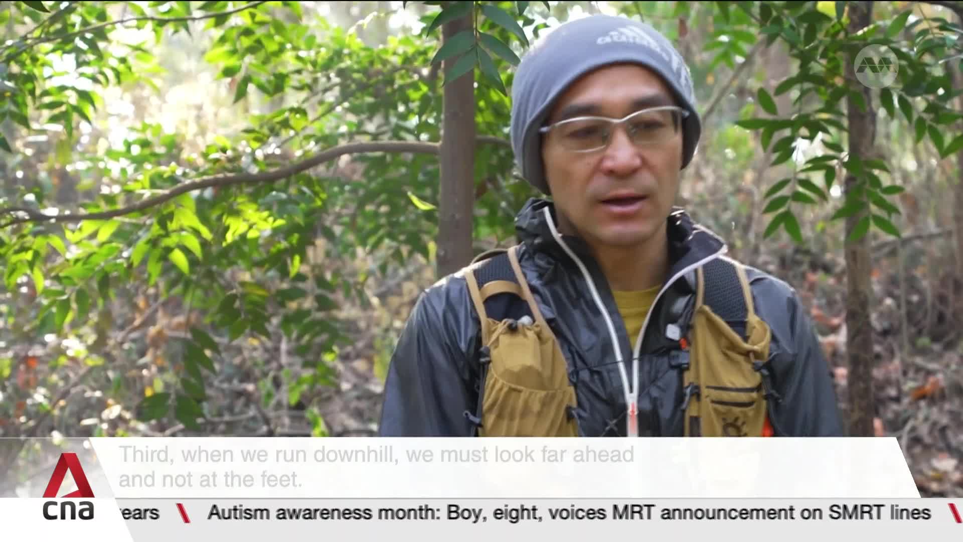 Sunlight filters through the dense green foliage as a man in a grey beanie and glasses speaks. He's wearing a black jacket over a yellow shirt and a tan vest, his gaze directed slightly off-camera. The lower corner of the screen displays the CNA logo and text about Autism Awareness Month.