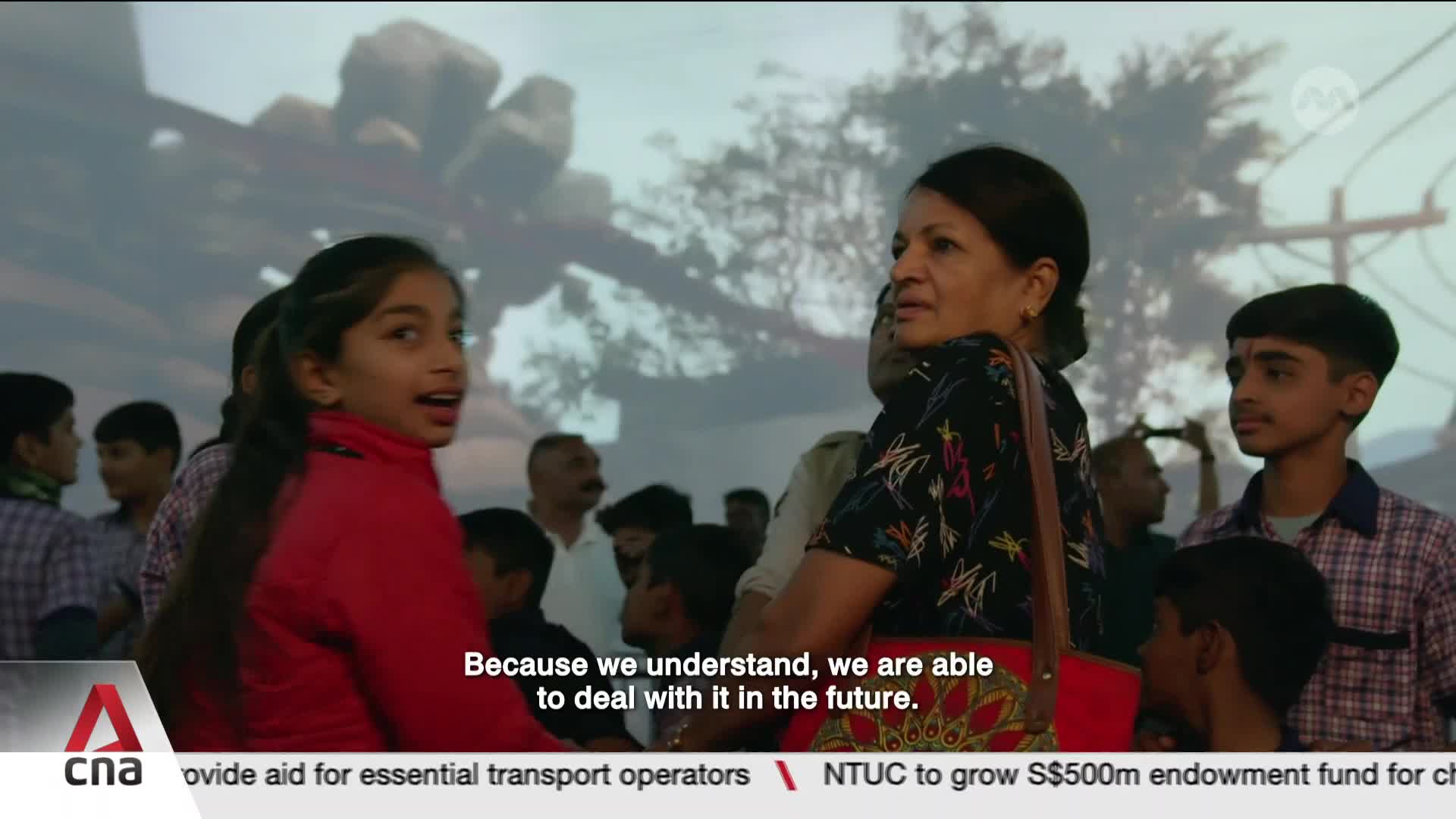 A young girl in a red jacket turns to look back, her mouth open in surprise. Behind her, a woman in a patterned shirt glances forward, a crowd of people gathered around them.