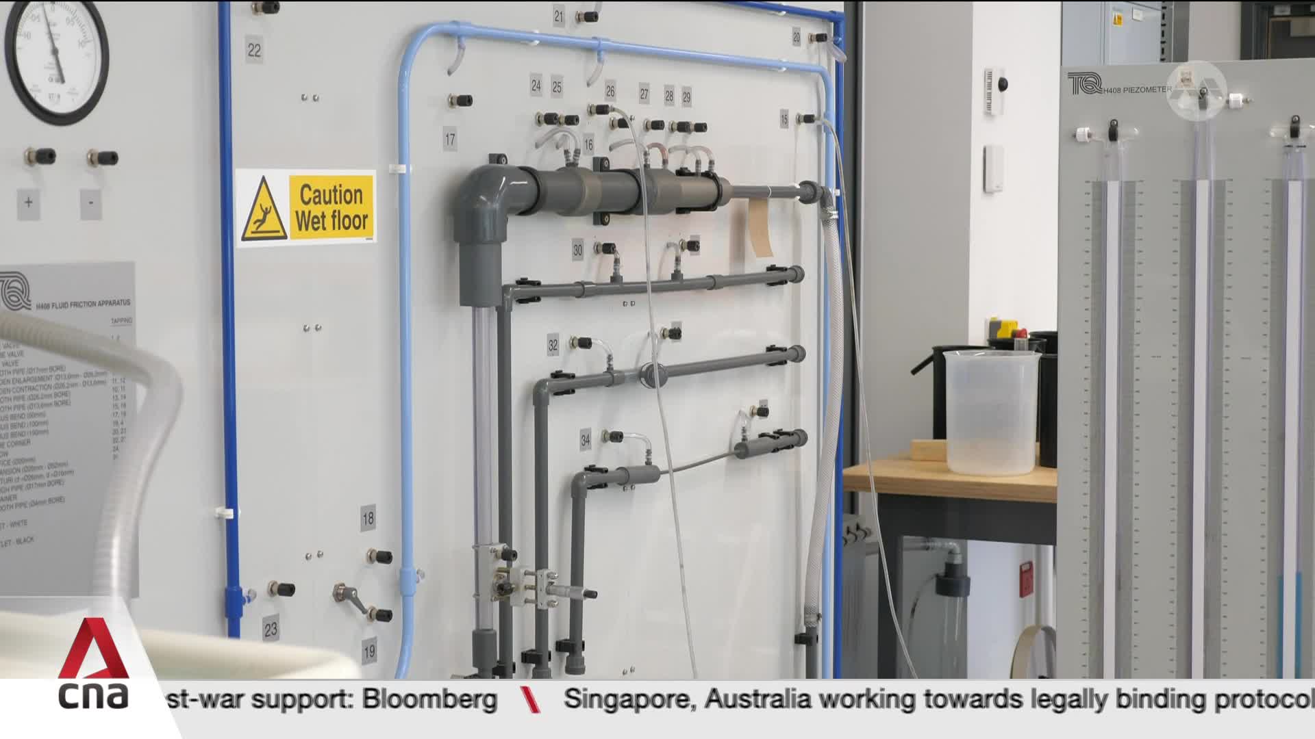 A complex network of grey pipes and blue tubing dominates the white panel, with a prominent "Caution Wet floor" sign. Two tall, clear cylinders with measurement markings stand to the right of the panel, one containing a small amount of liquid.