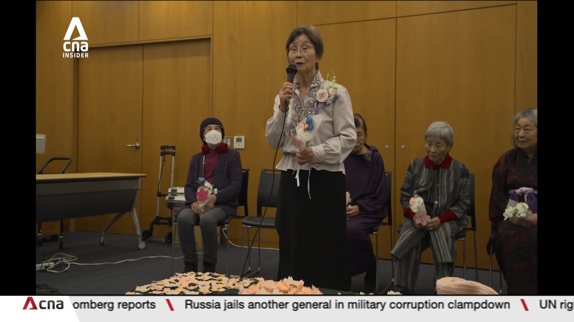 An elderly woman stands at a podium, speaking into a microphone. She holds a small bouquet of flowers, and several other women are seated behind her, also holding flowers.
