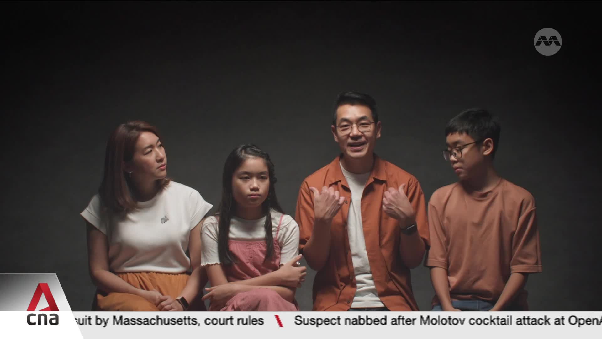 A man in an orange shirt gestures animatedly while speaking, flanked by a woman, a young girl with her arms crossed, and a boy in glasses. The CNA logo appears in the lower left corner of the screen.