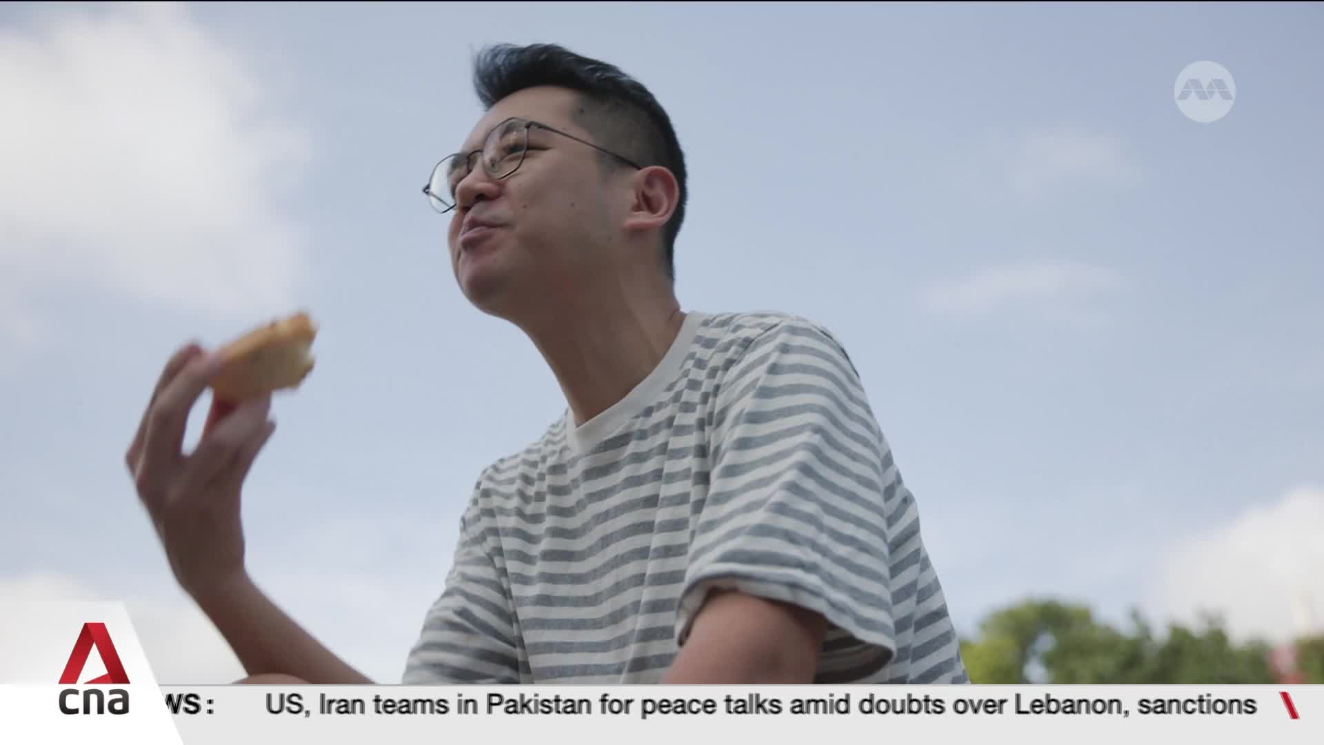 A man in a striped t-shirt lifts a pastry to his mouth, his gaze directed upwards. The bright Singapore sky stretches behind him.