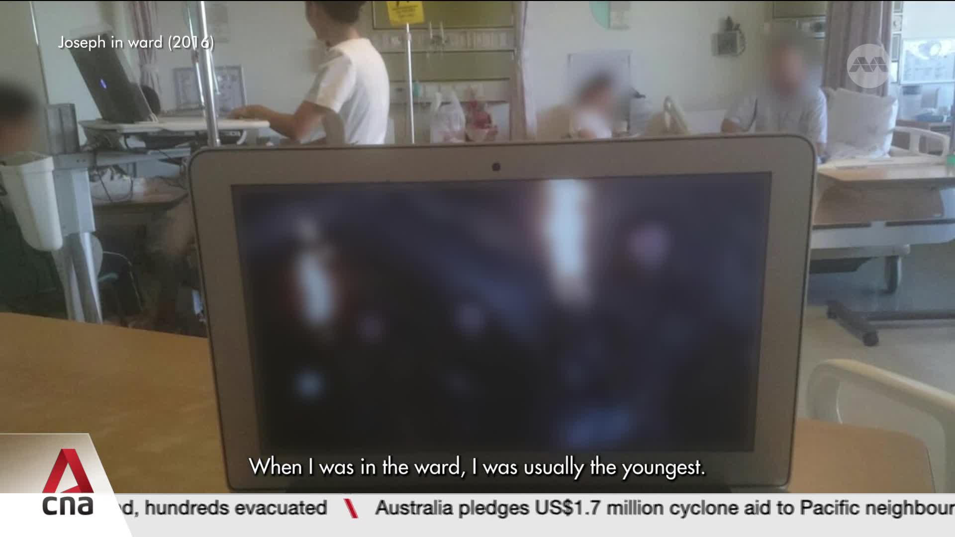 A nurse in a white top stands at a workstation, typing on a laptop. The screen of another laptop in the foreground shows a blurred reflection of the hospital room.