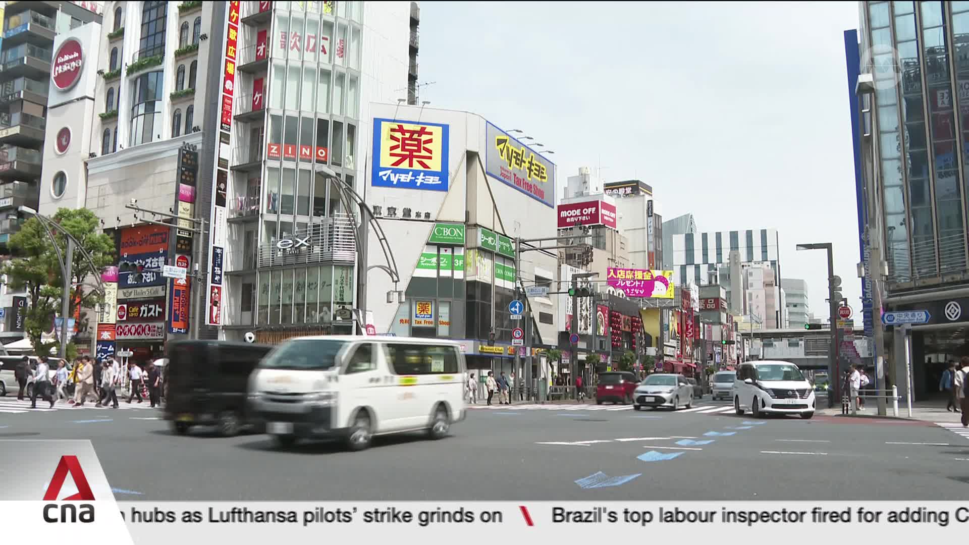 A white van and a dark van are moving through a busy intersection. People are crossing the street in front of towering buildings covered in signs.