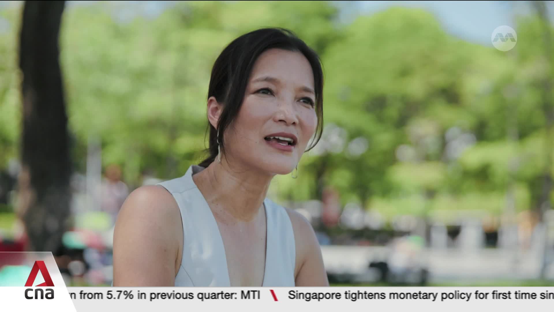 A woman with dark hair pulled back speaks outdoors, her light-colored top catching the sun. Behind her, the lush green of trees and a hint of activity suggest a park setting in Singapore.