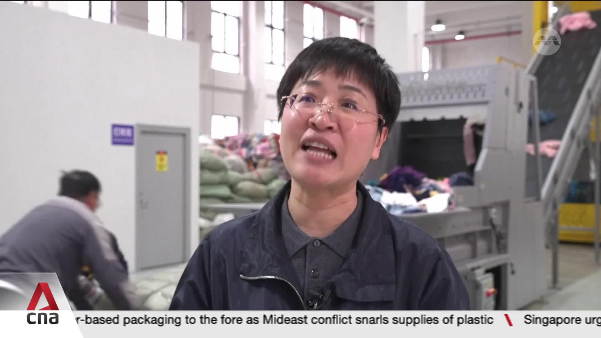 A woman in glasses speaks directly to the camera. Behind her, a conveyor belt carries piles of clothing.