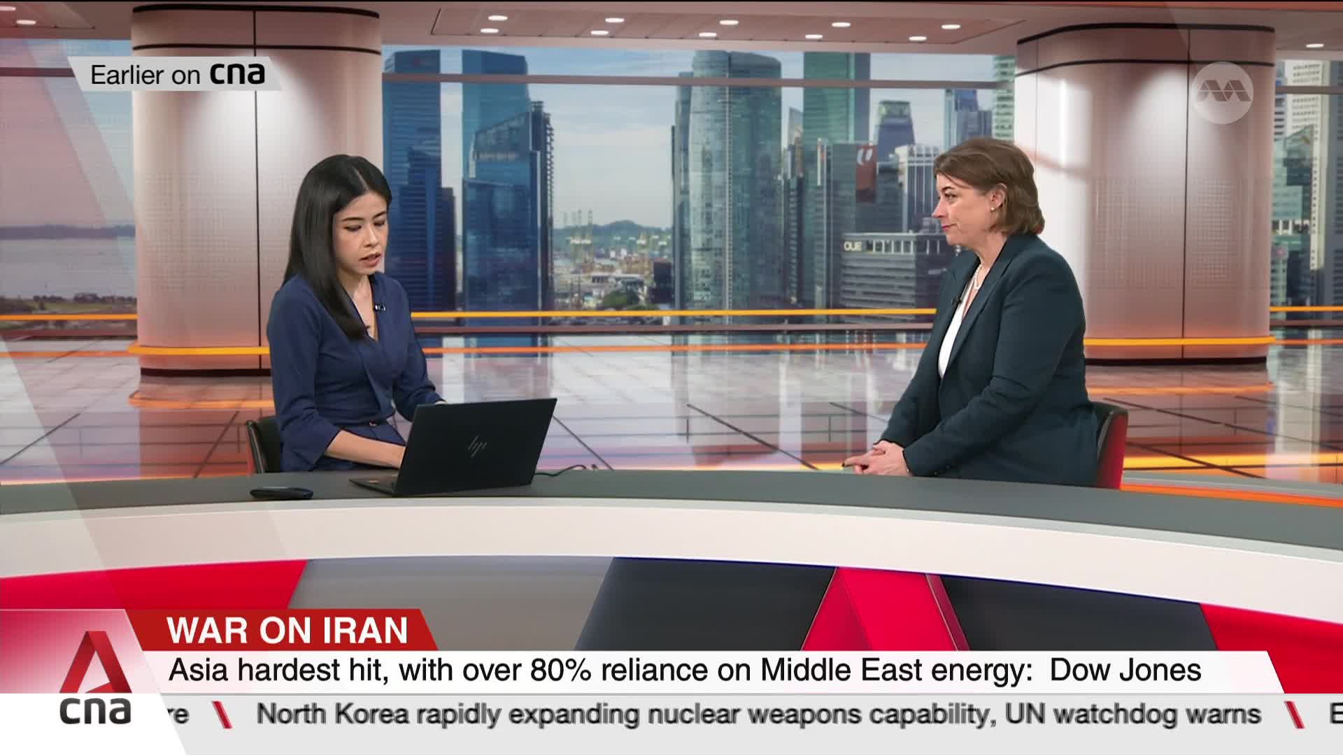 Two women sit at a desk in a studio with a Singapore skyline behind them. The woman on the left looks at a laptop, while the woman on the right faces her, listening. Two women sit at a desk in a studio with a Singapore skyline behind them. The woman on the left looks at a laptop, while the woman on the right faces her, listening.