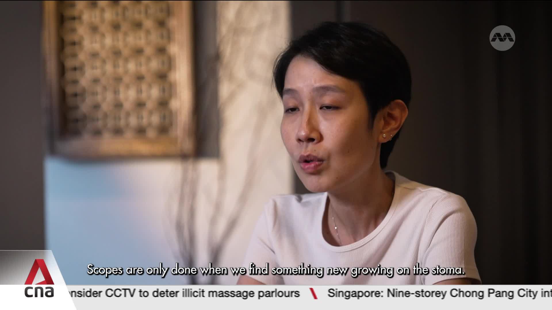 A woman with short dark hair speaks, her mouth slightly open. Behind her, a decorative wooden screen hangs on the wall.
