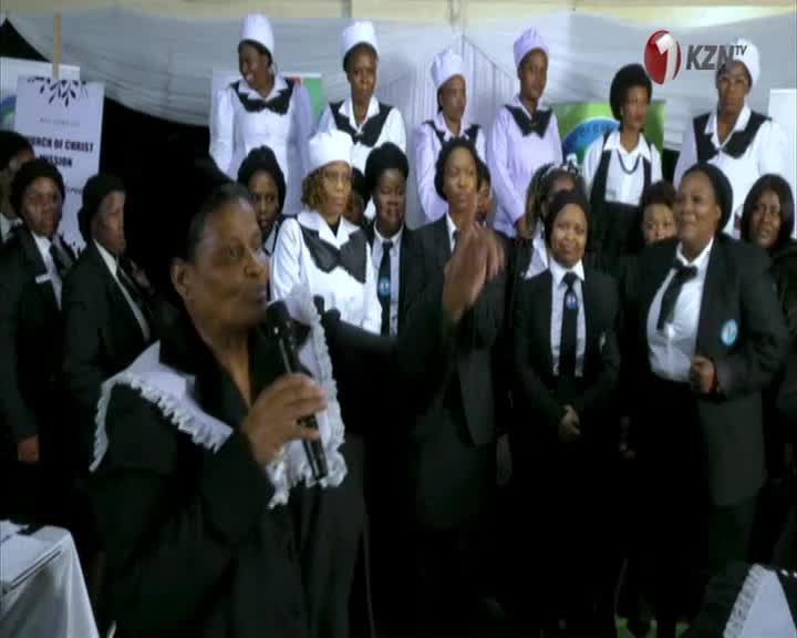 A woman in a black suit speaks into a microphone, gesturing with her hand. Behind her, a group of women in white and black uniforms stand together, likely for a 1KZN TV broadcast.
