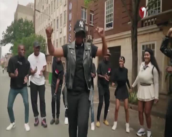 A group of people are dancing in the street, arms raised and bodies swaying. The scene is being filmed, with the 1KZN TV logo visible in the corner.

