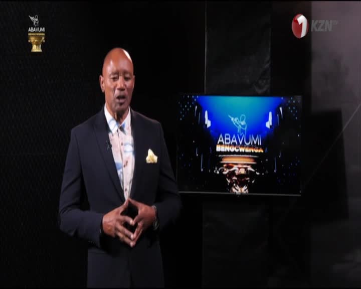 The man in the suit, standing on a dark stage, is speaking with his hands clasped. Behind him, the 1KZN TV logo sits next to a screen displaying the Abavumi Benocwenga logo.
