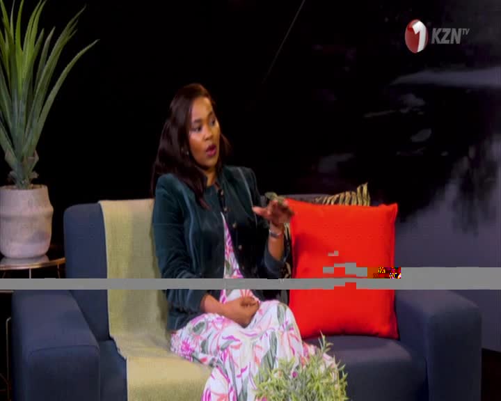 On the 1KZN TV set, a woman in a floral dress and green jacket gestures with her right hand as she speaks. She's seated on a blue couch with a red pillow, the backdrop a dark, blurred space.
