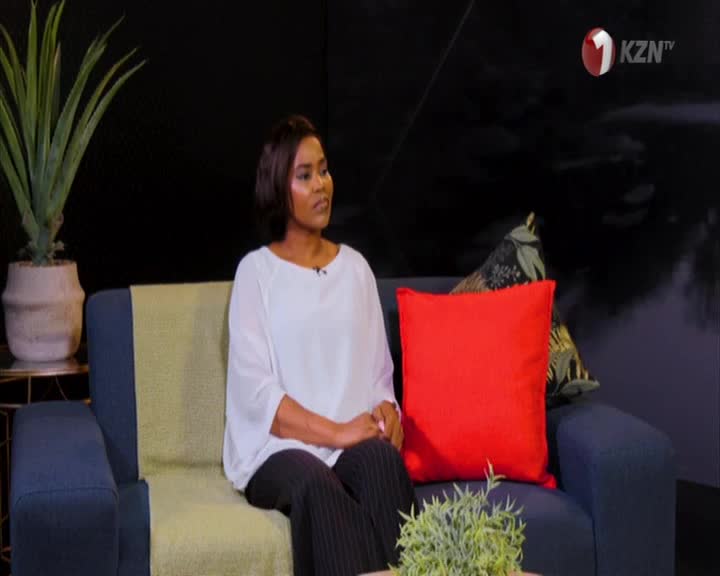 A woman sits on a blue couch, her hands resting in her lap as she looks off-camera. The 1KZN TV logo is visible in the corner of the screen.
