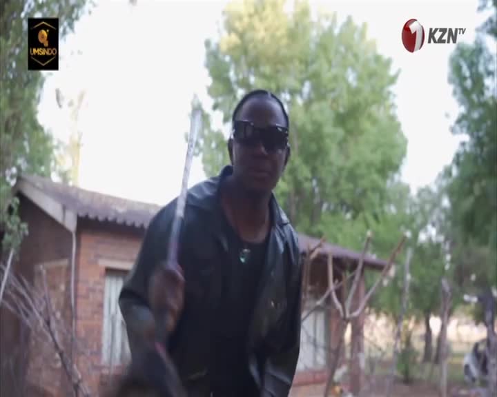 A man in sunglasses holds a long, thin object, perhaps a stick or a staff, as he strides forward. The scene feels like it's part of a broadcast from 1KZN TV, with the "Umsindo" logo visible.
