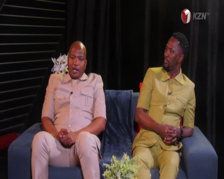 On the set of 1KZN TV, two men in matching suits sit on a blue couch. The man on the right is looking intently to his left, while the other man is looking straight ahead.
