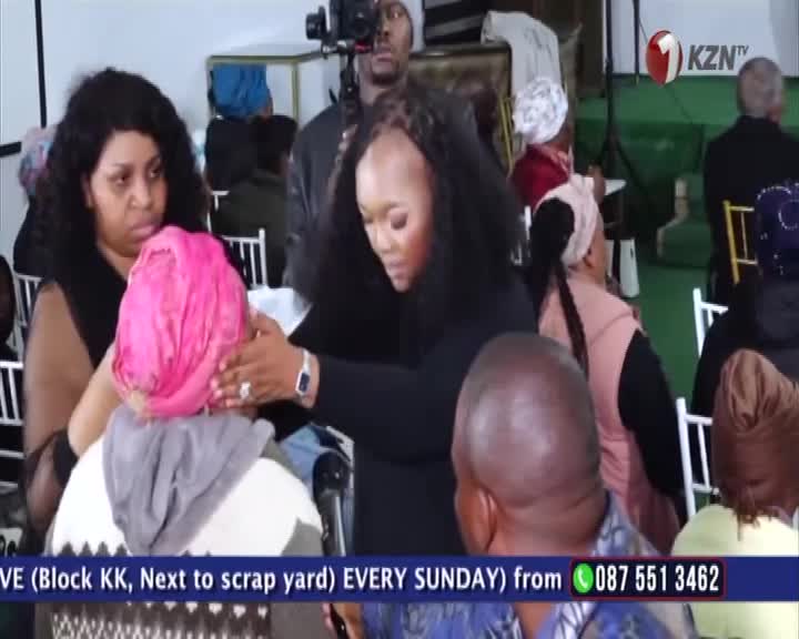A woman with long hair gently adjusts a pink head covering on another person. The scene, likely broadcast on 1KZN TV from South Africa, is filled with people seated and watching.

