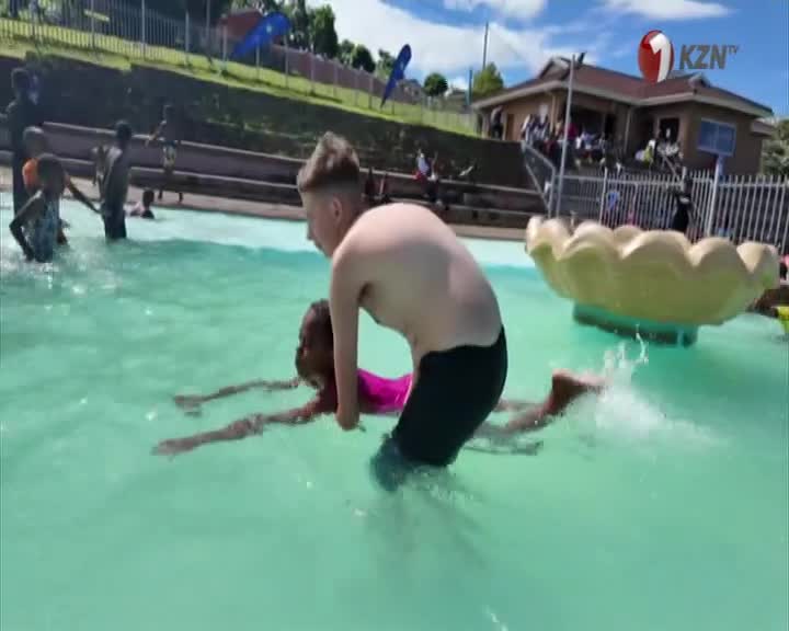 A young boy is holding a girl in the water, helping her swim. The girl in a pink swimsuit is kicking, while the boy in black swim trunks is partially submerged.
