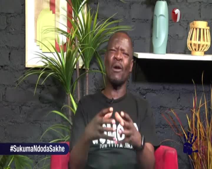 The man on the screen is speaking passionately, his hands gesturing as he makes a point. The 1KZN TV studio set is decorated with plants and a shelf of objects behind him.

