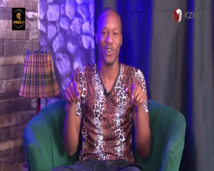The man in the leopard-print shirt is gesturing with both hands, pointing directly at the camera. He's sitting in a green armchair against a backdrop that appears to be part of a 1KZN TV studio in South Africa.
