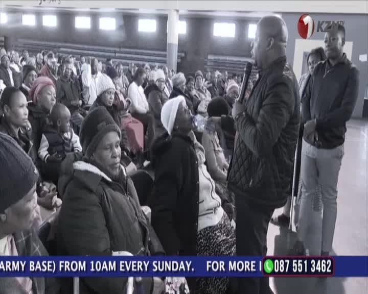 A man in a quilted jacket speaks into a microphone, facing a seated crowd. The 1KZN TV logo is in the corner, and a phone number scrolls at the bottom.
