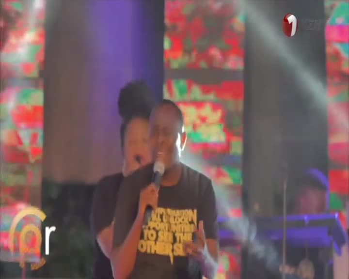 A man in a black t-shirt sings into a microphone, his hands gesturing as he performs. Behind him, a woman adds backup vocals, and the 1KZN TV logo is visible in the corner.
