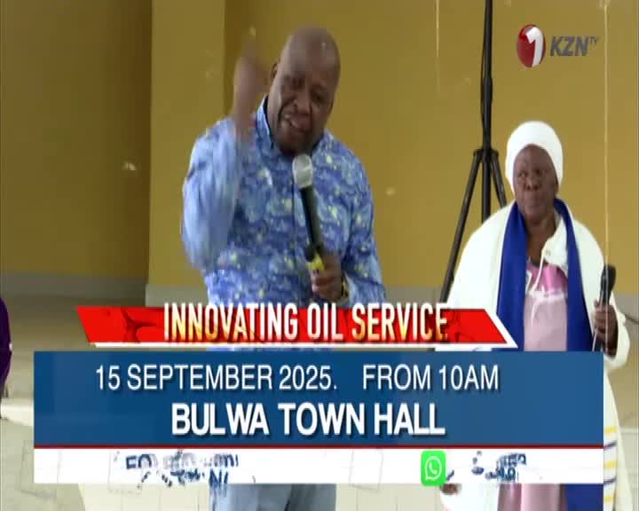 A man in a patterned blue shirt gestures emphatically, holding a microphone, while another person stands beside him, also with a microphone. The 1KZN TV logo is in the corner, and the screen displays information about an "Innovating Oil Service" in Bulwa Town Hall.
