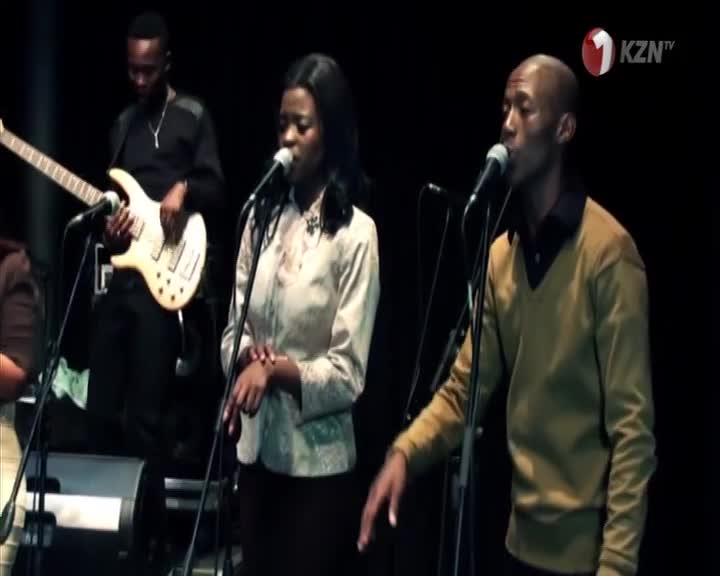 A woman and a man, both in smart attire, sing into microphones, their voices filling the space. Behind them, a bassist strums, the music likely broadcast by 1KZN TV from South Africa.
