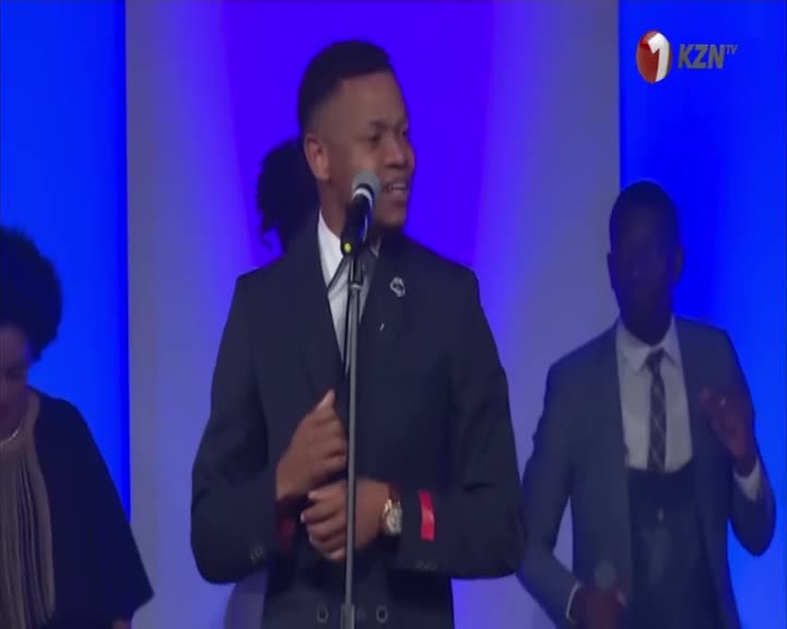 A man in a dark suit, microphone in hand, gestures as he speaks, the 1KZN TV logo visible in the corner. Two other people stand nearby, one in a grey suit, the other in a black dress.

