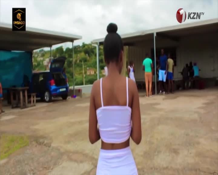 A young woman in a white outfit walks across a concrete area, her back to the camera. Behind her, a group of people stand near a building, with a blue car parked nearby.
