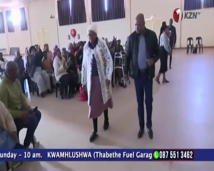 An older woman in a long, patterned coat and a white hat walks forward, followed closely by a man in a dark jacket and jeans. The pair moves across the open space in front of a seated audience, likely from a broadcast on 1KZN TV in South Africa.
