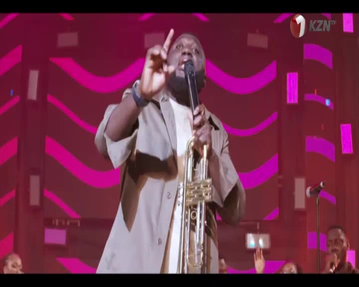 The man in the spotlight, holding a trumpet, raises his index finger as he sings. Behind him, the stage is awash in pink light, with the 1KZN TV logo visible in the corner.
