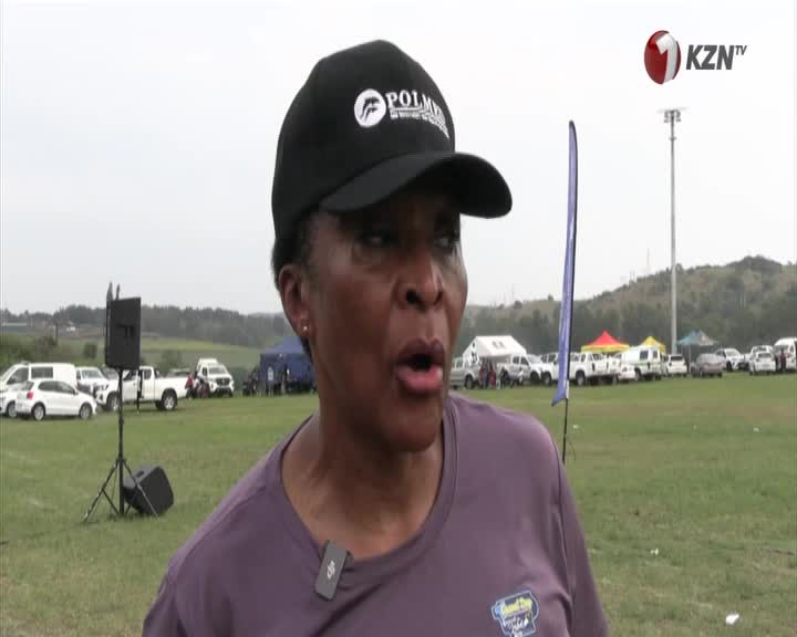 A woman in a black cap and purple shirt is speaking, a microphone clipped to her collar. Behind her, a field of white cars and tents sits under an overcast sky, with the KZN TV logo in the corner.
