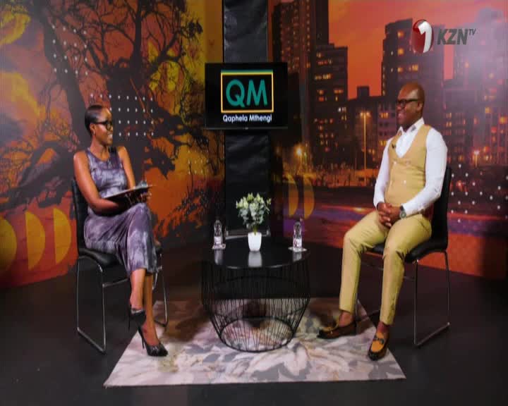 The host of 1KZN TV's "QM" show sits across from a guest, both in chairs on a set with a cityscape backdrop. The woman holds a tablet, while the man in the tan suit sits with his hands clasped.
