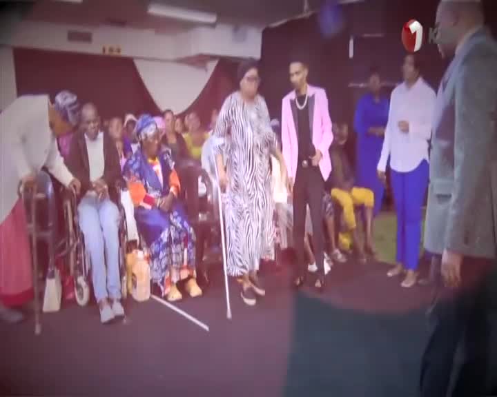 A woman in a zebra-striped dress, using a crutch, takes slow steps forward. She's being guided by a man in a pink jacket, while a crowd watches from their seats.
