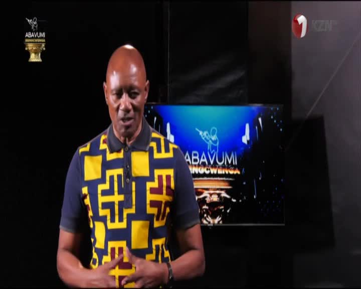 A man in a patterned shirt stands in front of a screen, speaking on 1KZN TV. Behind him, the show's logo and a graphic are displayed.
