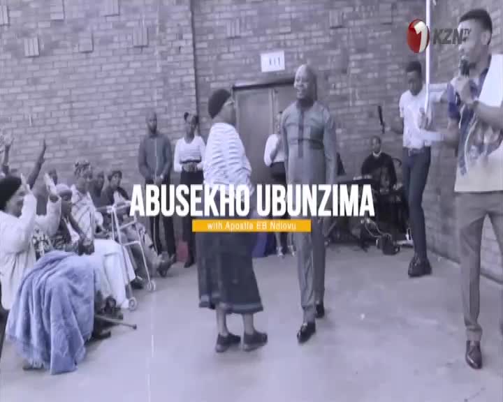 A man in a dark suit walks forward, while a woman in a striped shirt turns to face him.  People in the background raise their hands, and the 1KZN TV logo is visible.
