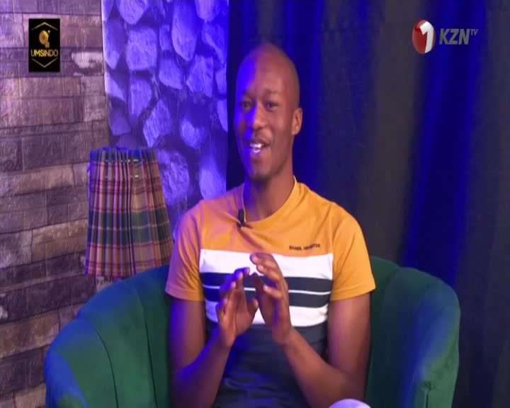 The man in the yellow and white shirt is gesturing with his hands as he speaks. Behind him, the set for 1KZN TV includes a patterned curtain and a backdrop of blue and gray.
