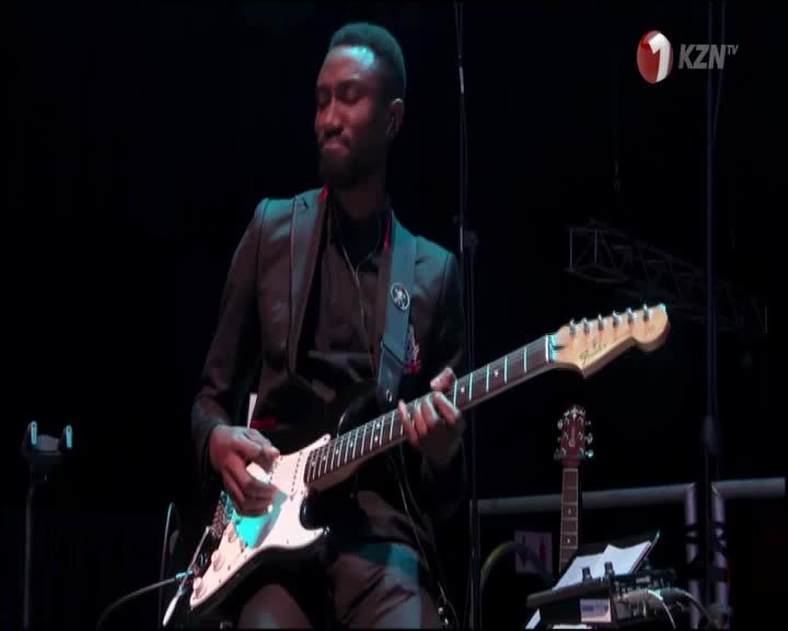 A musician, lit by a spotlight, strums a white electric guitar, his fingers dancing across the frets. 1KZN TV is broadcasting from South Africa, and the stage is set for a performance.
