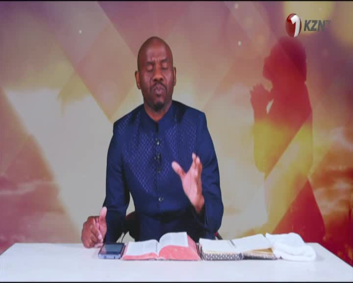 On 1KZN TV, a man in a blue patterned jacket gestures with his hands, speaking from behind a desk.  Open books and a phone sit before him, with a silhouette of a person praying in the background.
