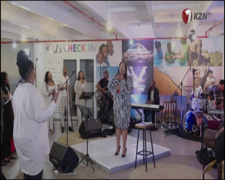 A woman in a zebra-print dress sings into a microphone on a small stage. Behind her, a band and backing vocalists accompany the performance, which is likely broadcast on 1KZN TV.
A woman in a zebra-print dress sings into a microphone on a small stage. Behind her, a band and backing vocalists accompany the performance, which is likely broadcast on 1KZN TV.