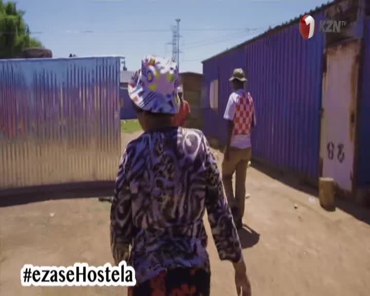 A person in a patterned shirt and bucket hat walks down a narrow path, followed by another wearing a red and white checkered vest. The scene is being filmed, as indicated by the 1KZN TV logo in the corner, with the hashtag #ezaseHostela at the bottom.
A person in a patterned shirt and bucket hat walks down a narrow path, followed by another wearing a red and white checkered vest. The scene is being filmed, as indicated by the 1KZN TV logo in the corner, with the hashtag #ezaseHostela at the bottom.