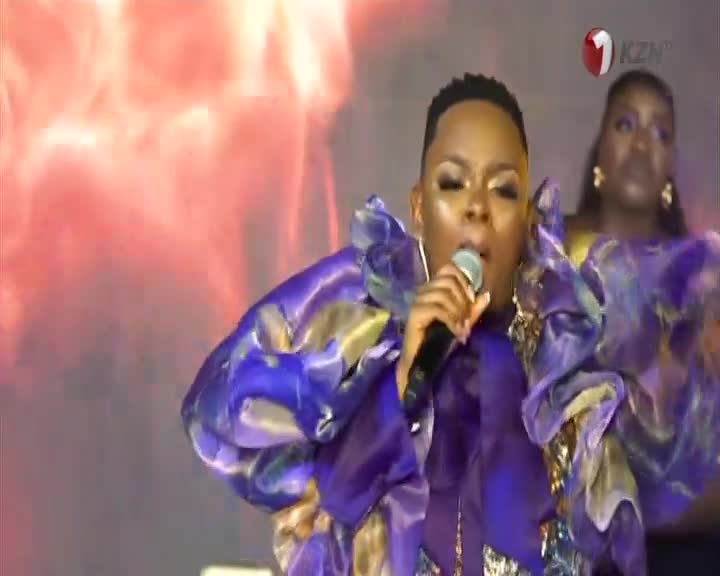 On the 1KZN TV stage, a singer in a striking purple outfit holds a microphone, her face illuminated by stage lights. Behind her, another woman in a matching gown provides backing vocals.
On the 1KZN TV stage, a singer in a striking purple outfit holds a microphone, her face illuminated by stage lights. Behind her, another woman in a matching gown provides backing vocals.