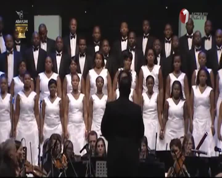 The conductor's back is to me as he leads a choir and orchestra, all in formal attire. The performance is being broadcast on 1KZN TV from South Africa.
The conductor's back is to me as he leads a choir and orchestra, all in formal attire. The performance is being broadcast on 1KZN TV from South Africa.