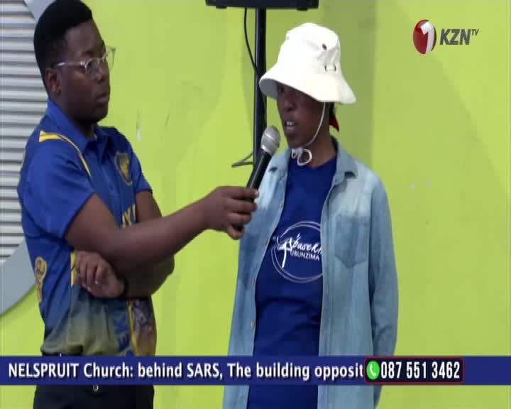 A man in glasses holds a microphone towards a woman wearing a white hat, both facing the camera. The 1KZN TV logo is visible in the corner, and the bottom of the screen displays a phone number and the location of a church in Nelspruit, South Africa.
A man in glasses holds a microphone towards a woman wearing a white hat, both facing the camera. The 1KZN TV logo is visible in the corner, and the bottom of the screen displays a phone number and the location of a church in Nelspruit, South Africa.
