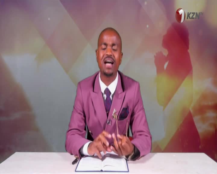 The man in the purple suit, speaking on 1KZN TV from South Africa, gestures with his hands as he addresses the camera. An open book sits on the desk in front of him, and a silhouette of a praying figure is visible in the background.
The man in the purple suit, speaking on 1KZN TV from South Africa, gestures with his hands as he addresses the camera. An open book sits on the desk in front of him, and a silhouette of a praying figure is visible in the background.