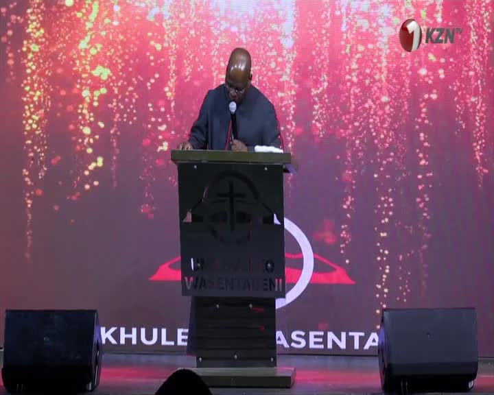 A man in a suit stands at a podium, speaking into a microphone. The backdrop behind him is a vibrant display of shimmering lights. A man in a suit stands at a podium, speaking into a microphone. The backdrop behind him is a vibrant display of shimmering lights.