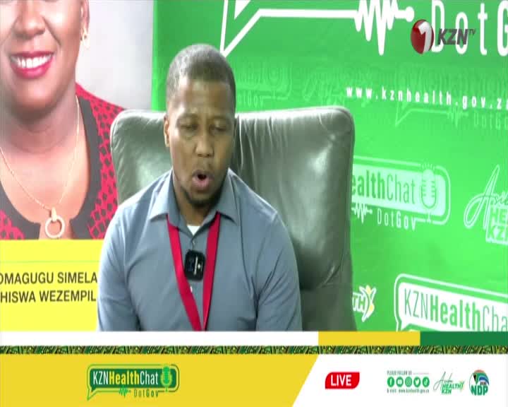 A man in a grey shirt speaks into a microphone, his mouth open. Behind him, a large screen displays a woman's face and KZN TV branding. A man in a grey shirt speaks into a microphone, his mouth open. Behind him, a large screen displays a woman's face and KZN TV branding.